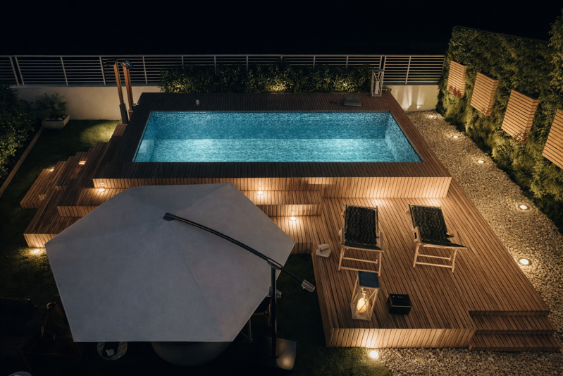 pazio esterno con piscina a Reggio Calabria – Vista aerea notturna della piscina sopraelevata con deck in legno e illuminazione integrata